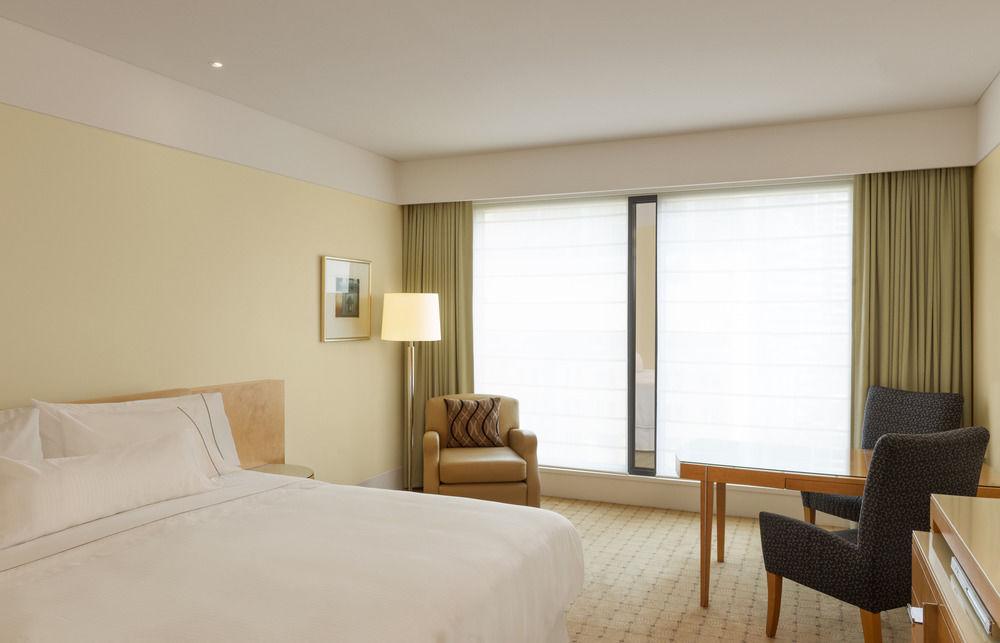 The Fullerton Hotel Sydney Eksteriør bilde The photo shows a modern hotel room. The decor features soft, neutral colors, creating a calm and inviting atmosphere. There is a large bed with crisp white linens against one wall. A bedside lamp stands on a small table next to the bed. 

Near the w
