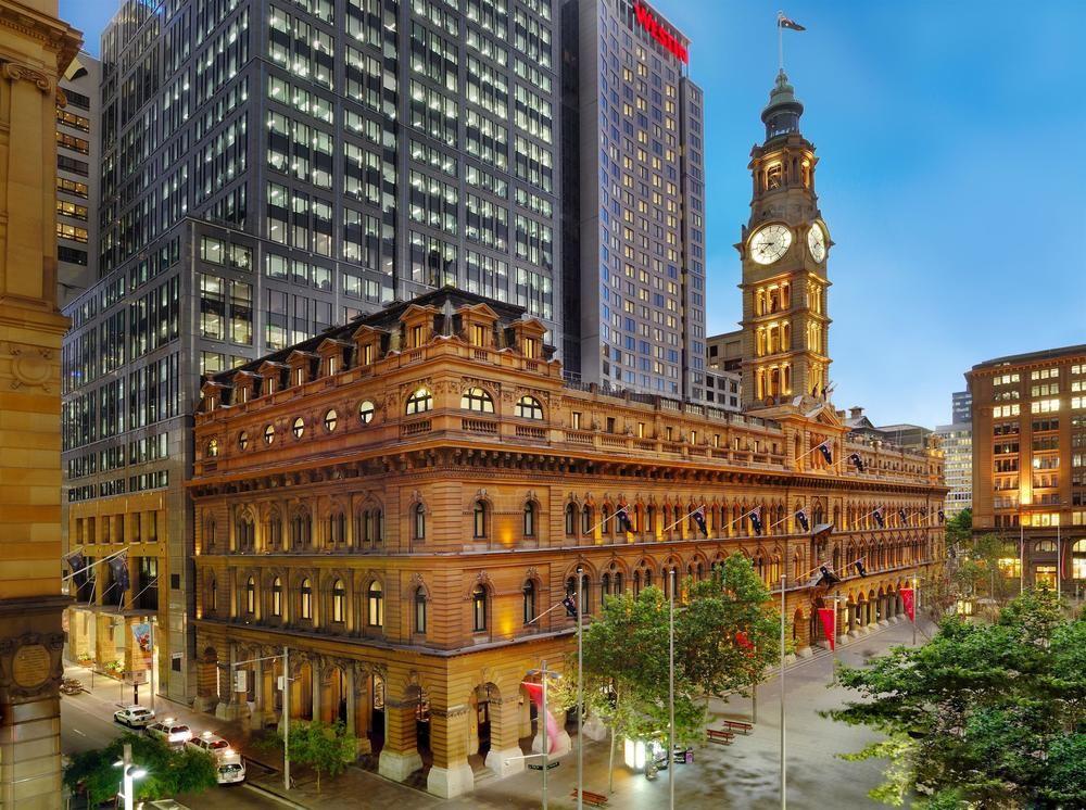 The Fullerton Hotel Sydney Eksteriør bilde The image depicts a contrasting architectural scene featuring a historic building alongside modern skyscrapers. The foreground shows a grand, ornate structure with decorative elements, likely a former post office or civic building, distinguished by i