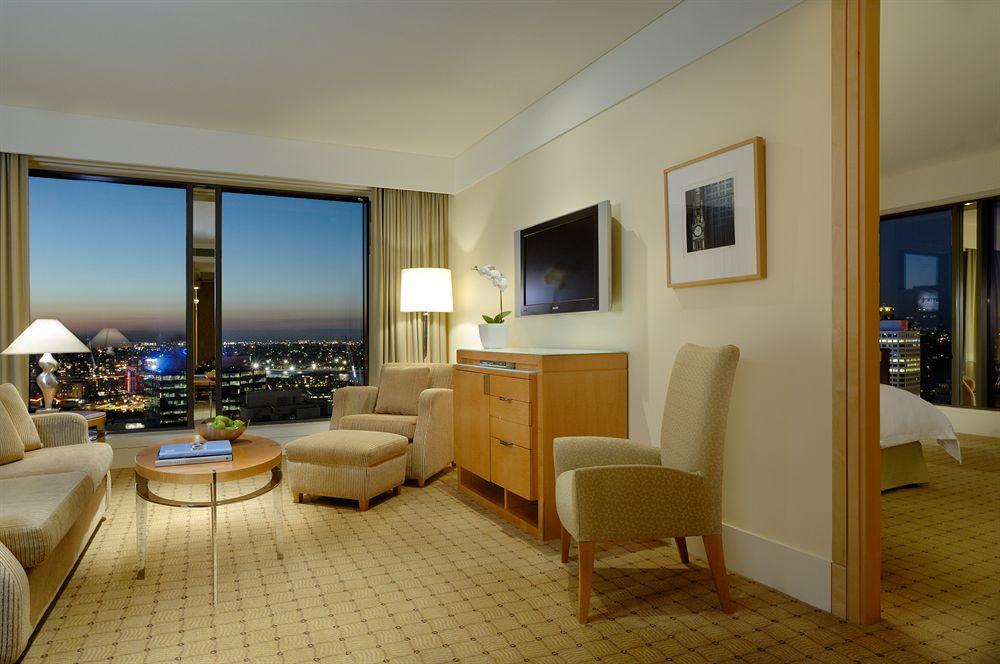 The Fullerton Hotel Sydney Eksteriør bilde The photo shows a hotel room with a modern and elegant design. There's a large window with a view of a city skyline, indicating it's likely at a higher floor. The room features a seating area with two light-colored chairs and a small round coffee tab