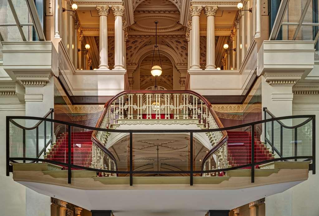 The Fullerton Hotel Sydney Interiør bilde The photograph depicts an elegant interior of a building, showcasing a grand staircase that curves gracefully between floors. The staircase features intricate railings and a plush red carpet that adds a touch of luxury. The space is adorned with tall