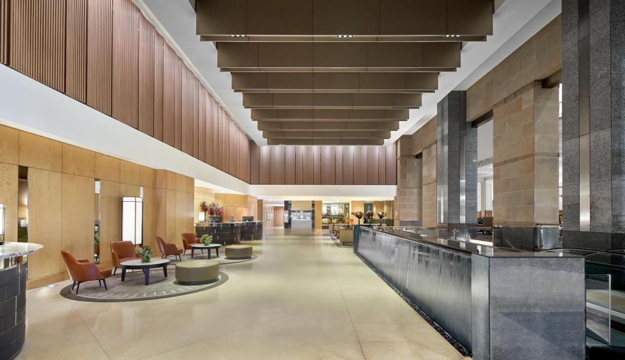 The Fullerton Hotel Sydney Eksteriør bilde The photo shows a spacious and modern hotel lobby. It features high ceilings with large, rectangular light fixtures hanging above. The walls are a combination of warm wood tones and stone, creating an elegant atmosphere. On one side, there are comfor
