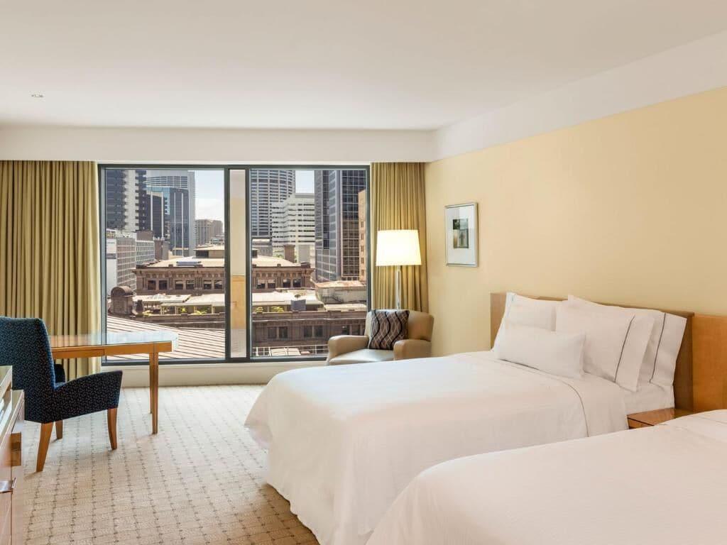 The Fullerton Hotel Sydney Eksteriør bilde The photo shows a hotel room with a modern design. There are two beds with white linens positioned in the center. A large window offers a view of a cityscape, featuring tall buildings outside. To the side, there is a comfortable armchair and a wooden