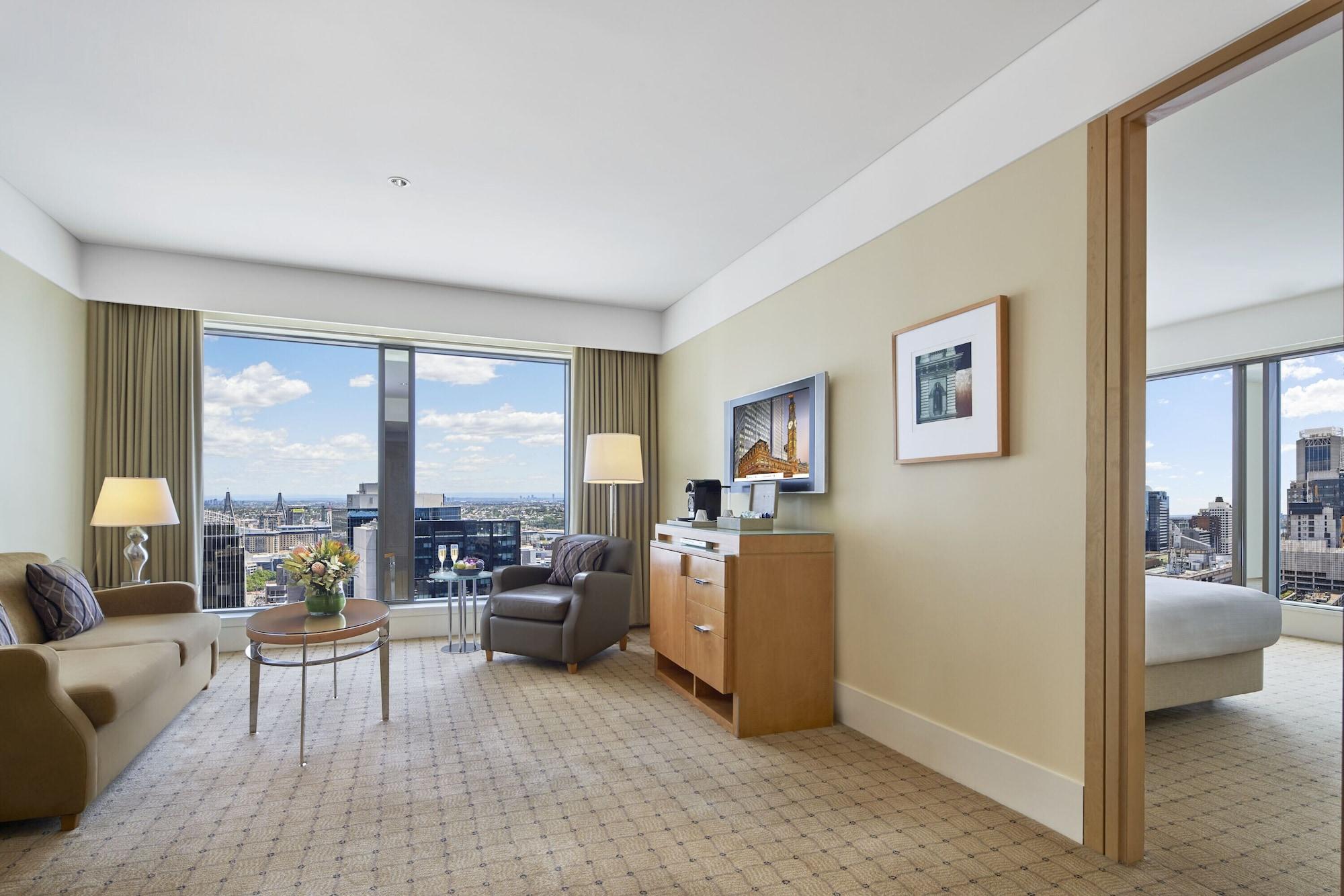 The Fullerton Hotel Sydney Eksteriør bilde The photo shows the interior of a hotel room. It features a spacious living area with large windows that offer a view of the city skyline. The room has beige walls and carpet, giving it a warm ambiance. There is a comfortable sofa, a coffee table, an