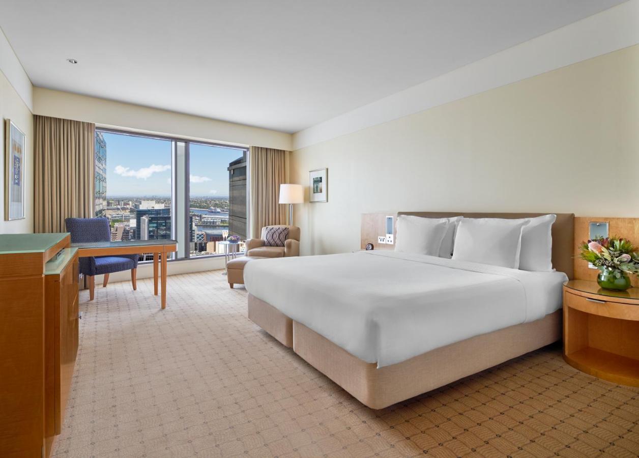 The Fullerton Hotel Sydney Eksteriør bilde The photo shows a hotel room featuring a large bed with white linens, set in a spacious area with light-colored walls. To one side, there is a comfortable chair and a small table, both positioned near a large window that allows for natural light and 