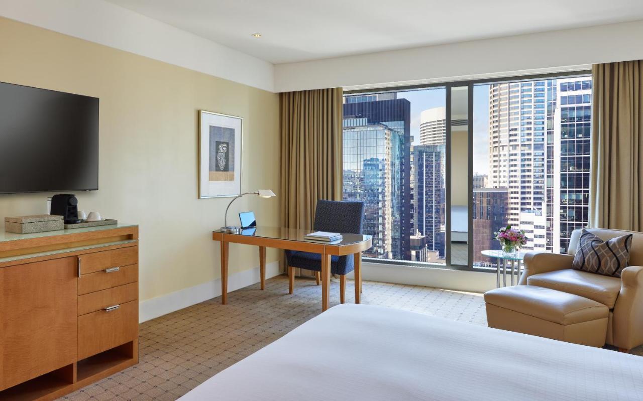 The Fullerton Hotel Sydney Eksteriør bilde The photo shows a modern hotel room with a cozy and contemporary design. There is a comfortable bed with white linens, a desk with a chair and a lamp positioned by a large window that offers a view of a cityscape filled with tall buildings. A flat-sc