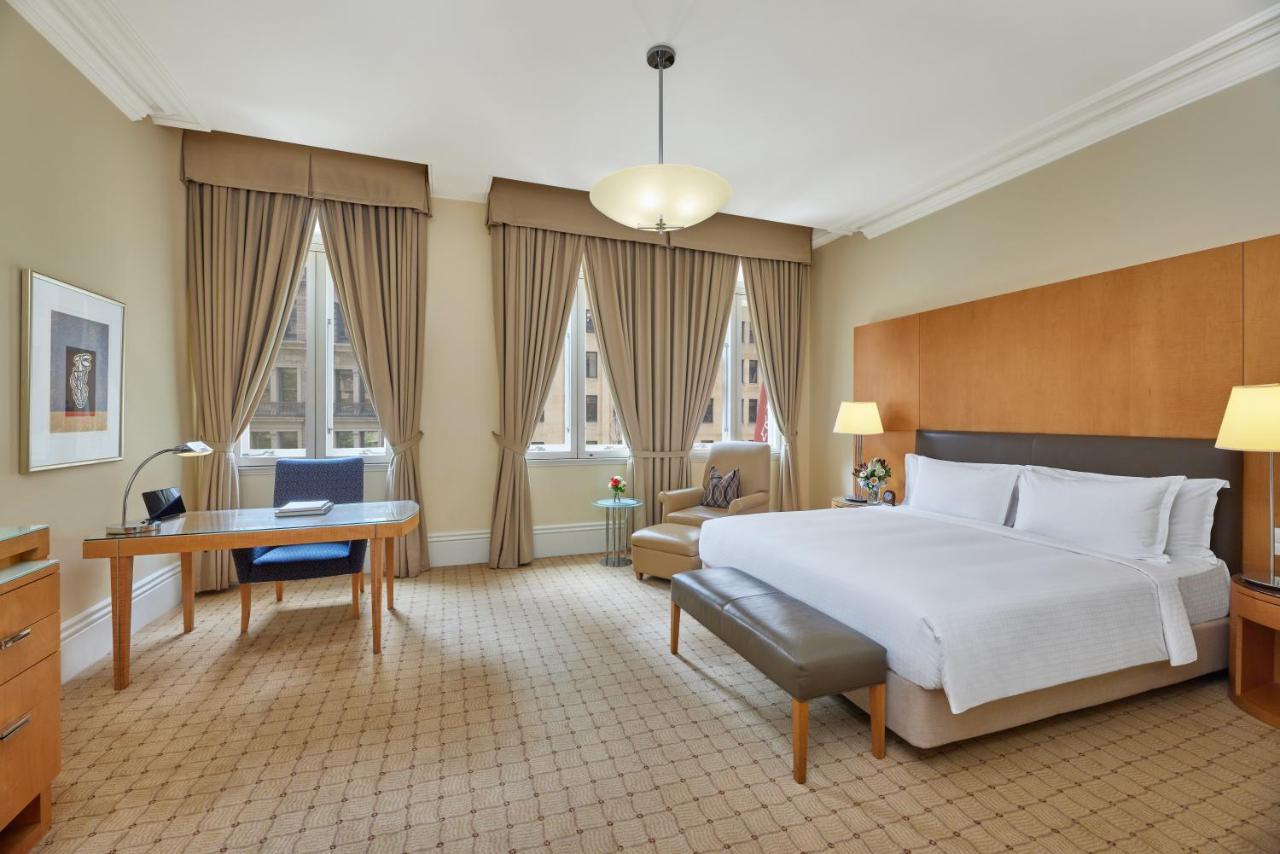 The Fullerton Hotel Sydney Eksteriør bilde The photo shows a spacious hotel room featuring a large bed with white linens. There is a modern bedside table and a small couch at the foot of the bed. A comfortable armchair is positioned nearby. Natural light enters through large windows adorned w