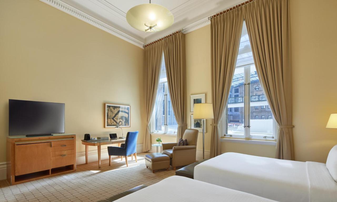 The Fullerton Hotel Sydney Eksteriør bilde The photo shows a hotel room featuring a light-colored interior with high ceilings. There are two beds positioned at the forefront of the image, with a small bedside table between them. To one side, a wooden dresser supports a flat-screen television.