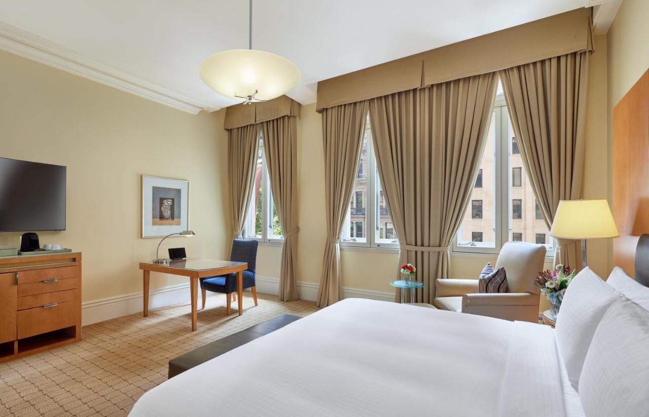 The Fullerton Hotel Sydney Eksteriør bilde The photo shows a well-furnished hotel room. It features a large bed with white linens, a desk with a chair, and a comfortable armchair beside it. The room has large windows draped with beige curtains, allowing natural light to enter. A flat-screen T