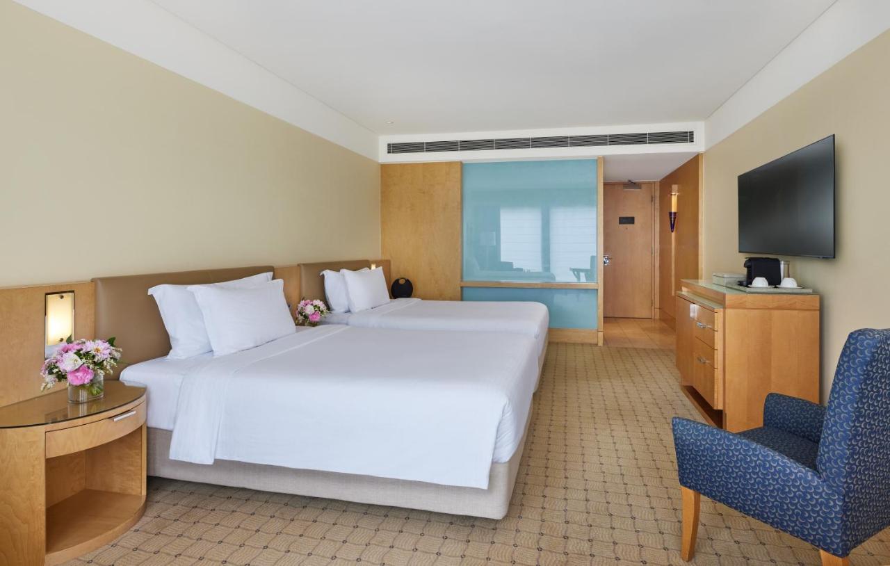 The Fullerton Hotel Sydney Eksteriør bilde The image shows a hotel room featuring two twin beds with white linens and decorative pillows. The walls are painted in a soft beige color, and there is wooden furniture, including a bedside table and a media console. A flat-screen TV is mounted on t