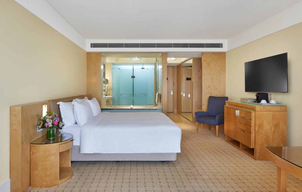 The Fullerton Hotel Sydney Eksteriør bilde The photo shows a modern hotel room. It features a large bed with white linens, a small bedside table with a vase of flowers, and a comfortable blue armchair in one corner. There is a wooden dresser and a flat-screen TV mounted on the wall. Behind th