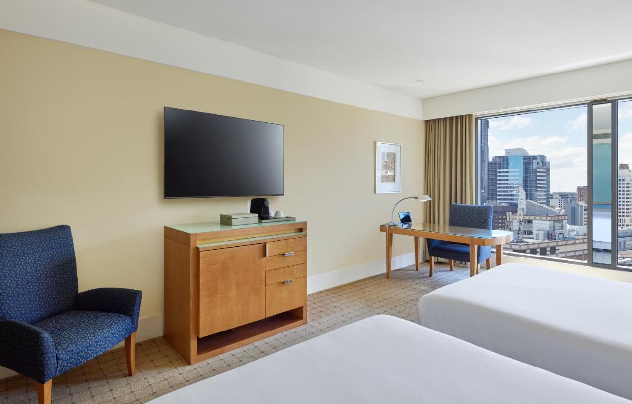 The Fullerton Hotel Sydney Eksteriør bilde The photo shows a modern hotel room featuring two beds with white linens. There's a wooden bedside table and a blue upholstered chair nearby. A television mounted on the wall is visible, alongside a wooden dresser with some items on top. Additionally