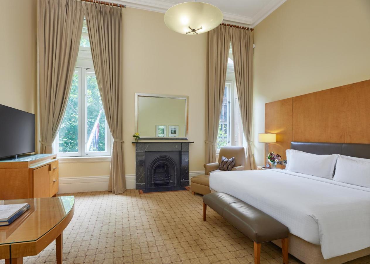 The Fullerton Hotel Sydney Eksteriør bilde The photo depicts an elegantly furnished hotel room. It features a large bed with white linens, a padded bench at the foot of the bed, and a comfortable armchair in one corner. There is a flat-screen TV on a wooden cabinet, and a stylish mirror above