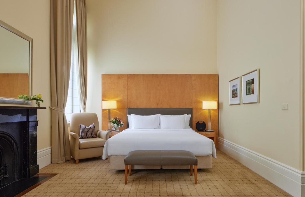The Fullerton Hotel Sydney Eksteriør bilde The photo shows a well-appointed hotel room. It features a large bed with white linens and a gray headboard, positioned against a wooden wall panel. On either side of the bed are stylish lamps. There is a comfortable armchair in the corner, accompani
