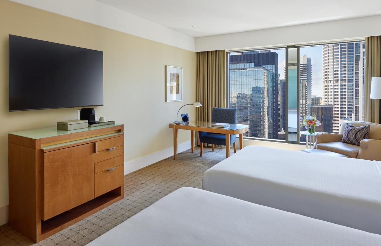 The Fullerton Hotel Sydney Eksteriør bilde The photo shows a hotel room featuring two beds with white bedding. There is a wooden furniture piece with a television mounted on the wall above it. To the side, there is a desk with a chair and a lamp, positioned near a large window that offers a v