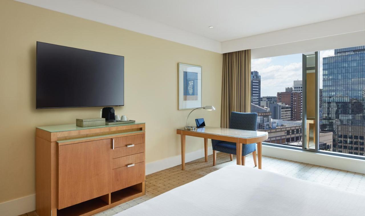 The Fullerton Hotel Sydney Eksteriør bilde The photo displays a modern hotel room with a clean and contemporary design. It features a large flat-screen television mounted on the wall. There is a wooden dresser with drawers beneath the TV. A small desk with a lamp and a chair is positioned nea