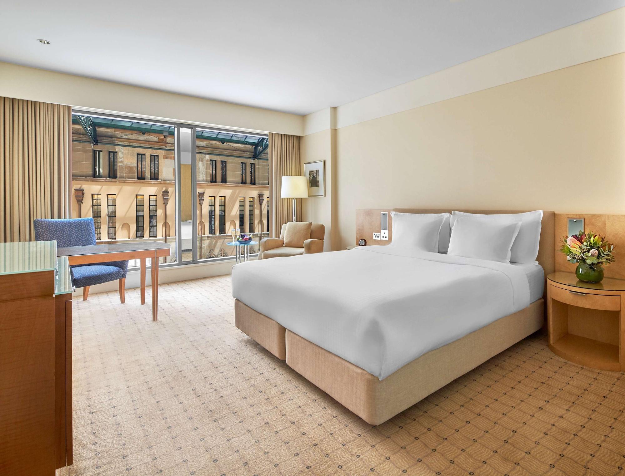 The Fullerton Hotel Sydney Eksteriør bilde The photo shows a hotel room featuring a large bed with white bedding at the center. On one side of the room, there is a light-colored bedside table with a lamp. Across from the bed is a wooden table accompanied by a blue chair, while a comfortable a
