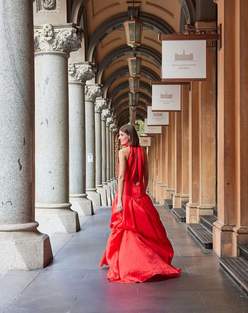 The Fullerton Hotel Sydney Eksteriør bilde The photo depicts a woman wearing an elegant red gown, standing in a covered archway lined with columns. She is turned slightly to look back over her shoulder, showcasing the flow and design of the dress. The environment features multiple arches and 