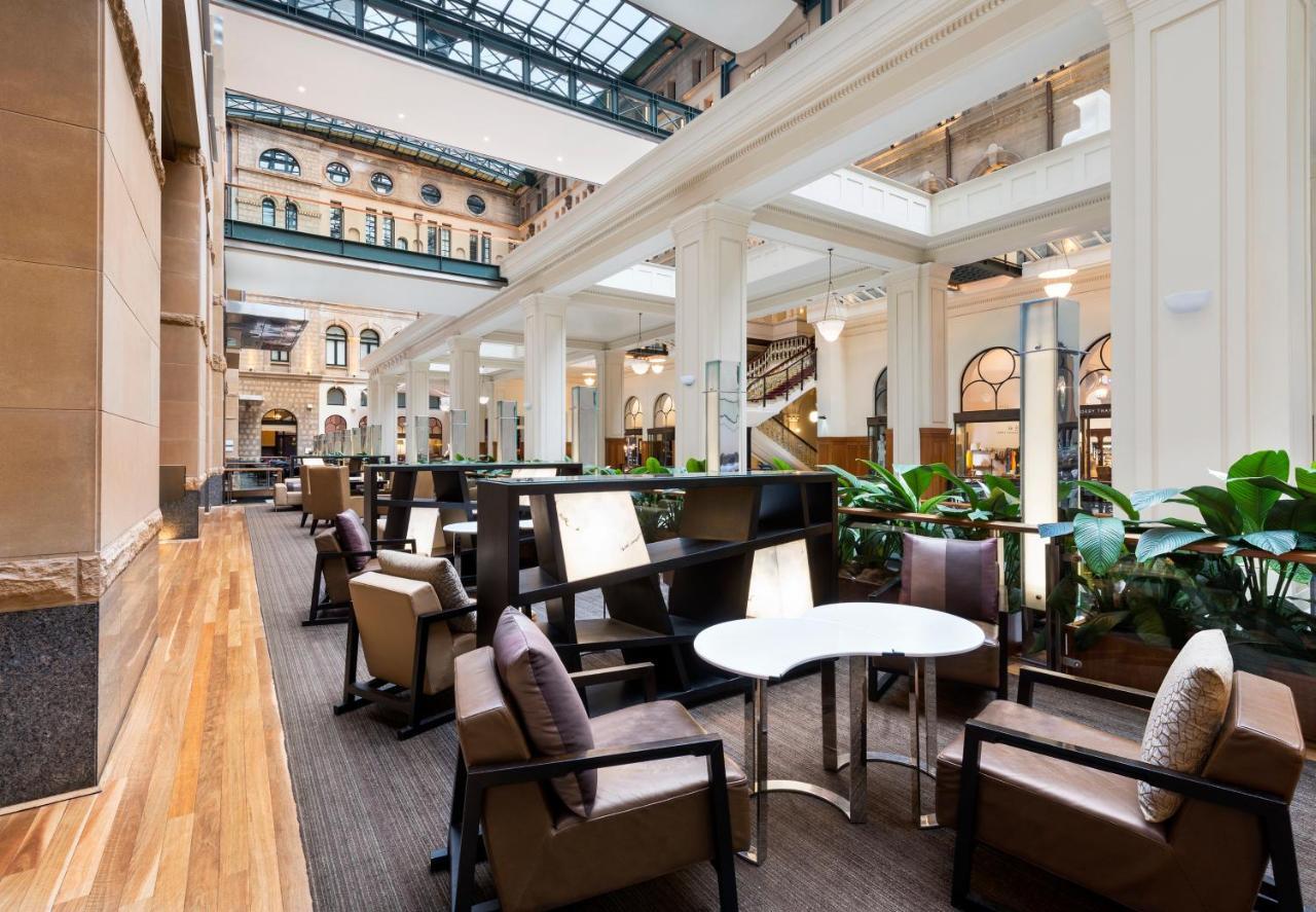 The Fullerton Hotel Sydney Eksteriør bilde The photo depicts a stylish, modern interior space, likely a hotel lobby or lounge area. It features tall ceilings with large windows that allow plenty of natural light to enter. The flooring is a combination of wood and carpet, creating a warm atmos